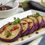 Tranches d'aubergines japonaises marinées, servies sur une assiette blanche avec une sauce épicée, garnies de coriandre et de graines de sésame. Une aubergine entière et un bol de sauce sont visibles en arrière-plan